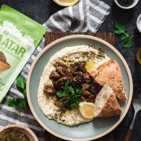 Ein Teller mit Hummus, gebratenem Lammfleisch, Pita-Brot, einer Zitronenscheibe und mit Zaatar bestreut, daneben eine Packung Nabali Zaatar.